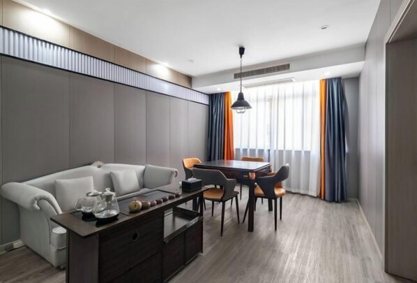 Une chambre d’hôtel moderne avec un canapé gris, une table basse en bois, une table à manger avec des chaises et une grande fenêtre avec des rideaux.