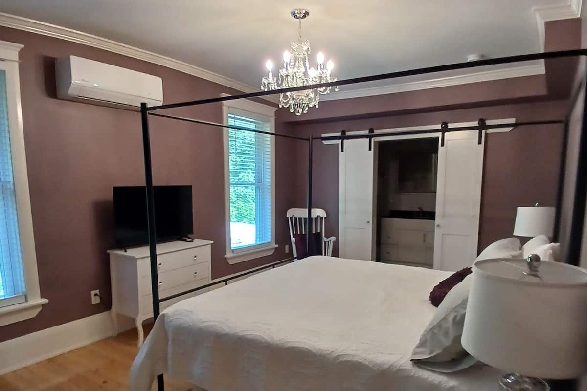 Un dormitorio con cama con dosel, una araña de techo, un televisor sobre un tocador y una ventana con persianas.