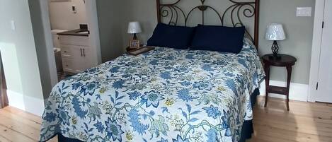Une chambre à coucher comprenant un lit, un ventilateur de plafond, une table de chevet avec une lampe et une porte donnant sur une salle de bain.