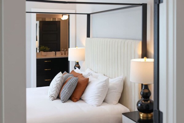 A four-poster bed with white linens and multiple pillows, two bedside lamps, and a view into a bathroom with a mirror and dark cabinetry.