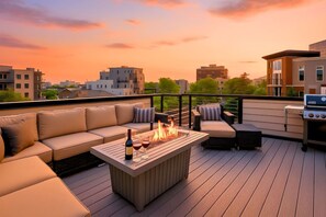 A rooftop deck with a wooden table, a built-in fire pit, outdoor seating, and a grill.