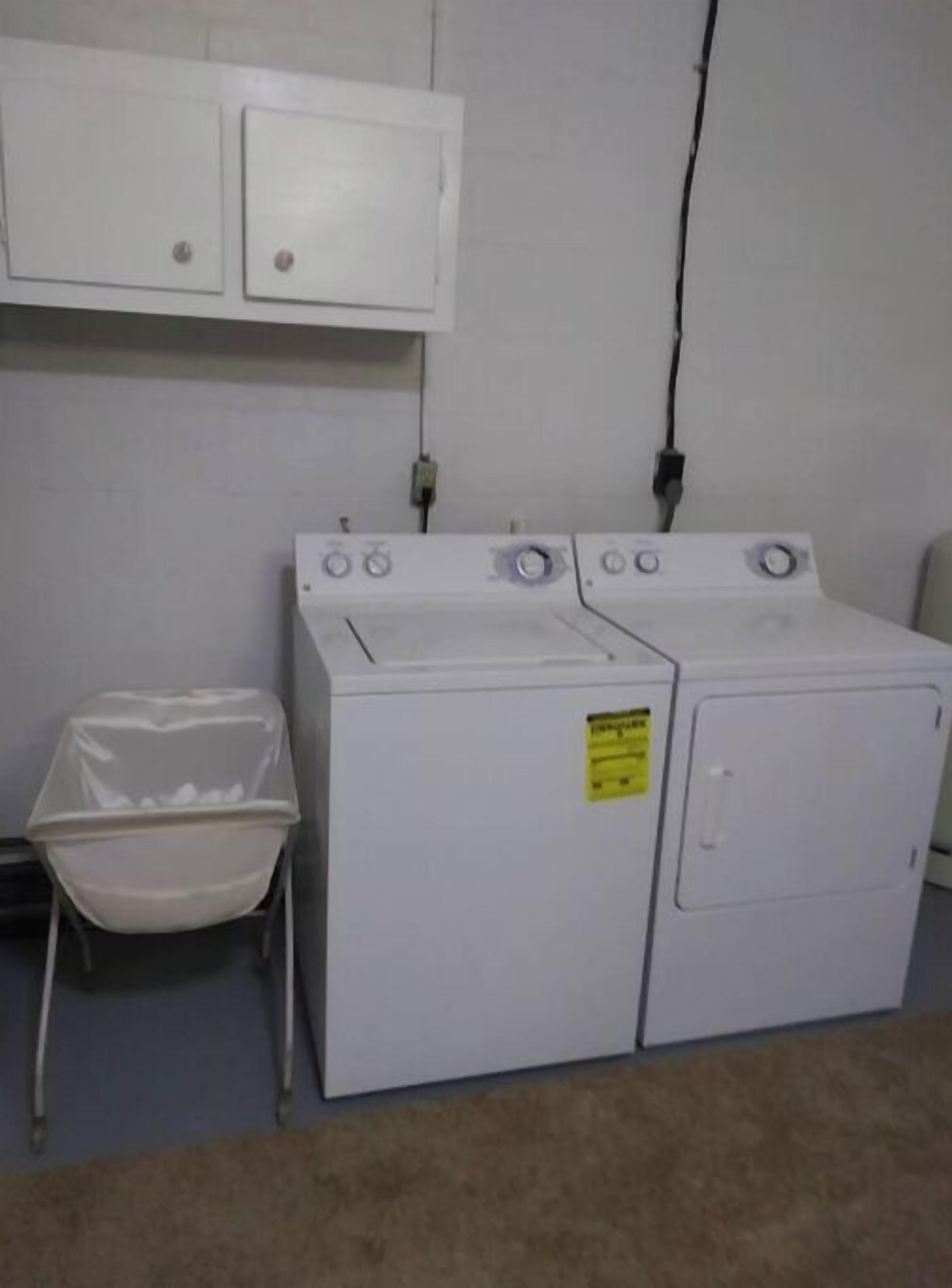 A laundry room with a front-loading washer and dryer, a small trash bin, and a wall-mounted electrical outlet.