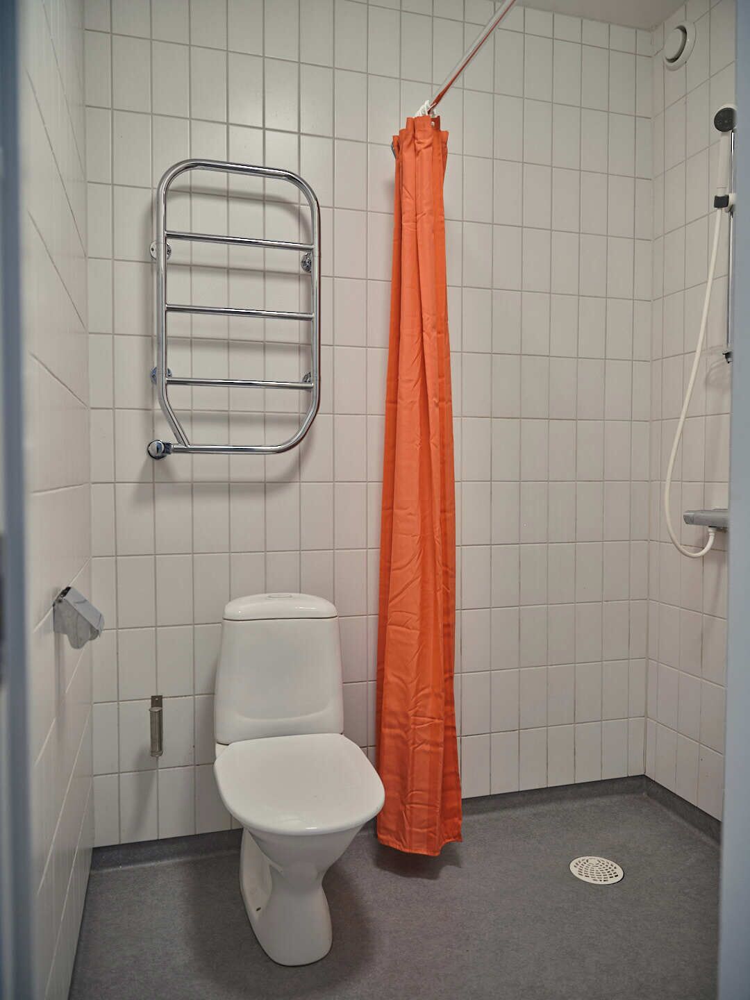 A small, tiled bathroom with a white toilet, a metal towel rack, and an orange shower curtain.