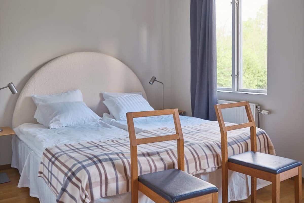 Une chambre à coucher avec un lit, deux chaises en bois, une table de chevet, une lampe et une fenêtre avec des rideaux.