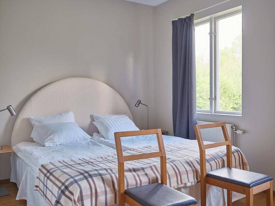 A bedroom with a bed, two wooden chairs, a nightstand, a lamp, and a window with curtains.