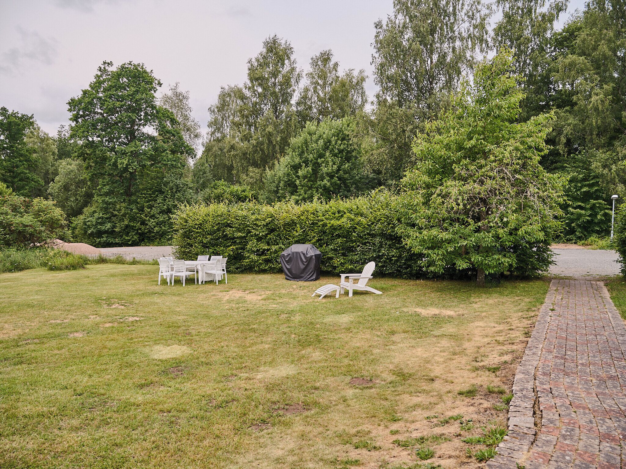 Une cour arrière verdoyante avec un chemin en briques, des meubles d’extérieur blancs et un barbecue.