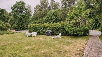 Une cour arrière verdoyante avec un chemin en briques, des meubles d’extérieur blancs et un barbecue.