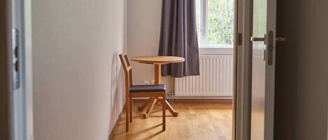 A hallway with wooden flooring, a table with a chair, a window with curtains, and a ceiling light fixture.