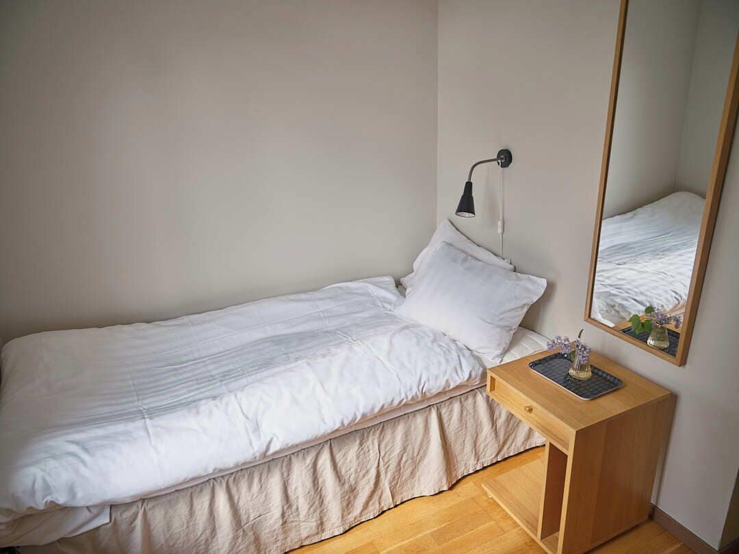 A single bed with a white duvet, a bedside table with a lamp, and a mirror reflecting the bed.