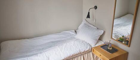 A single bed with a white duvet, a bedside table with a lamp, and a mirror reflecting the bed.