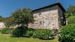 A stone building with a tiled roof, surrounded by well-maintained greenery and a clear blue sky. - Casa do Lagar (Cabeceiras de Basto)