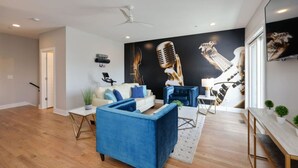 A modern living room with a blue sofa, a white sofa, and a blue armchair. There is a large wall mural of musical instruments. - Spacious West End | Rooftop Skyline Retreat (Nashville)