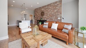 A modern living room with a brick accent wall, a leather sofa, a wooden coffee table, and a white armchair. - Spacious West End | Rooftop Skyline Retreat (Nashville)