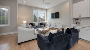 A modern living room with a sofa set, a coffee table, and a TV. There is a kitchen area with cabinets and a microwave.