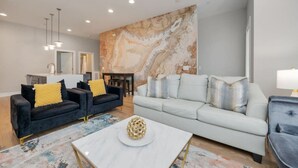 A modern living room with a large abstract painting, two sofas, a coffee table, and a dining area with a kitchen in the background.