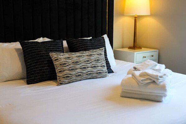 Una cama bien hecha con almohadones estampados, una toalla doblada y una lámpara de noche sobre una mesita.