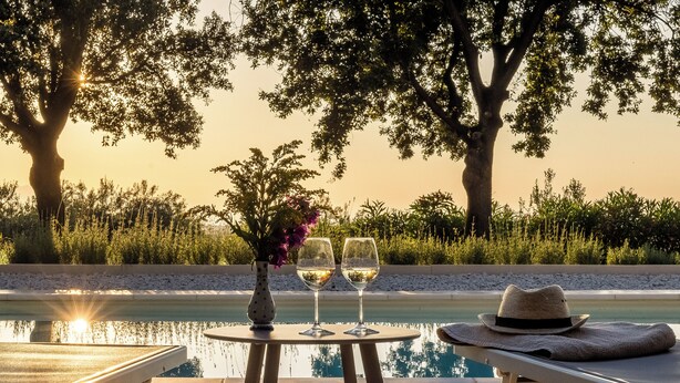 Un espace aménagé au bord de la piscine, avec deux chaises longues, une table sur laquelle reposent deux verres à vin et un vase de fleurs, et un chapeau posé sur la chaise de droite.