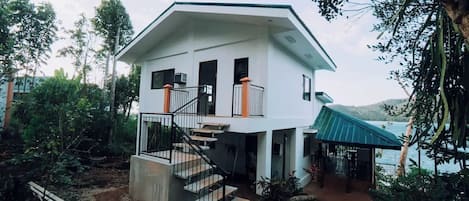 A two-story house with a staircase leading to the upper floor, surrounded by trees and a body of water.