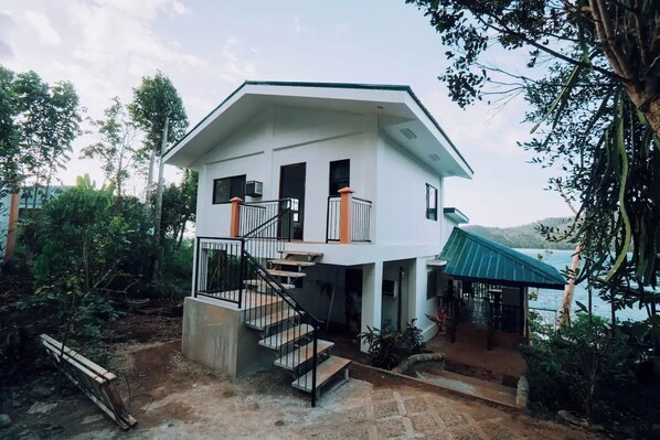 A two-story house with a staircase leading to the upper floor, surrounded by trees and a body of water.