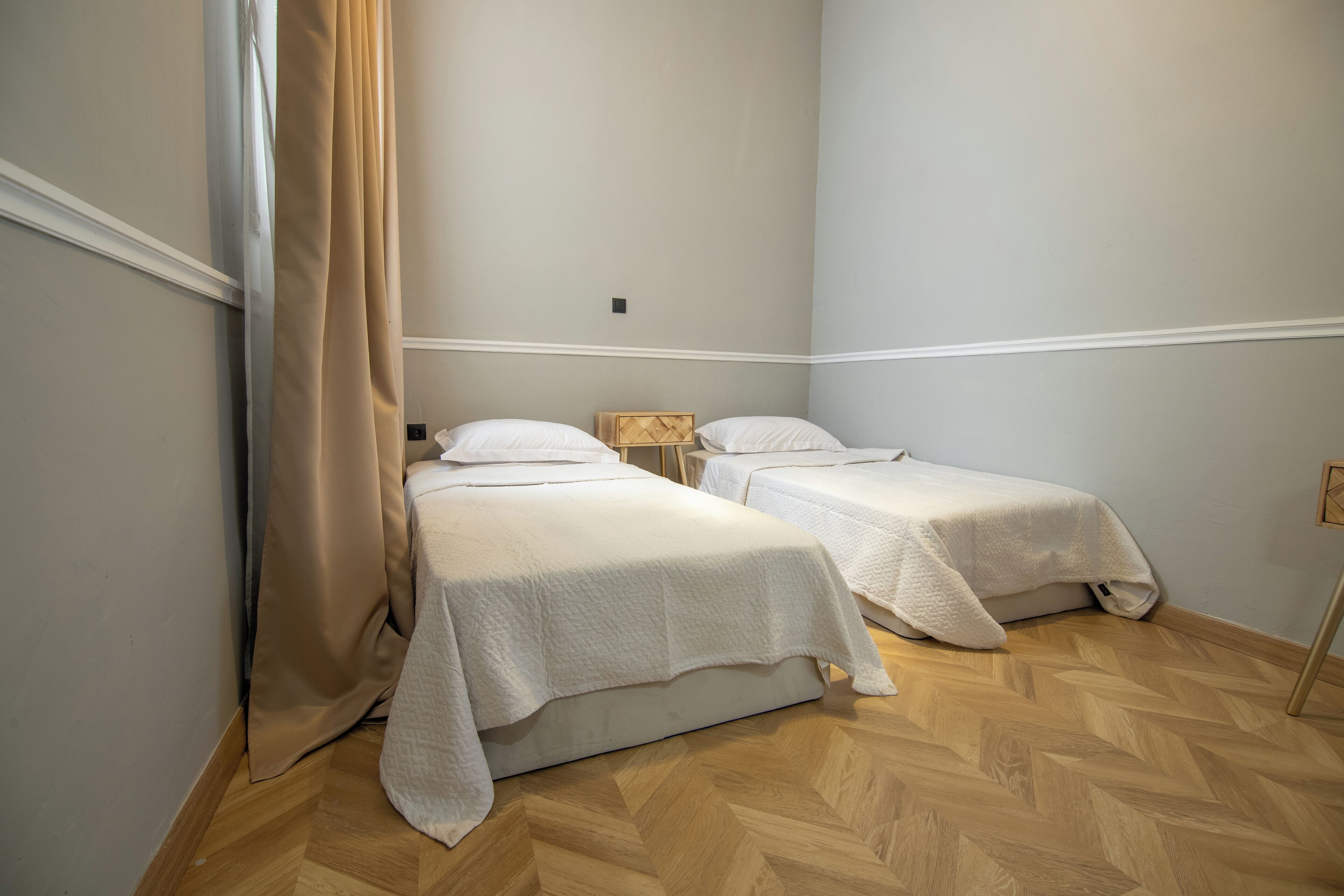 Une chambre avec deux lits simples, un parquet en bois disposé en point de Hongrie et un rideau beige.