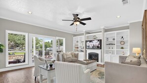 A spacious living room with a fireplace, a ceiling fan, and a large window overlooking a garden. - The Spot at Lake Granbury (Granbury)