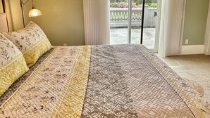 Une chambre avec un grand lit, une table de chevet, une lampe et une vue sur un balcon avec des rideaux.