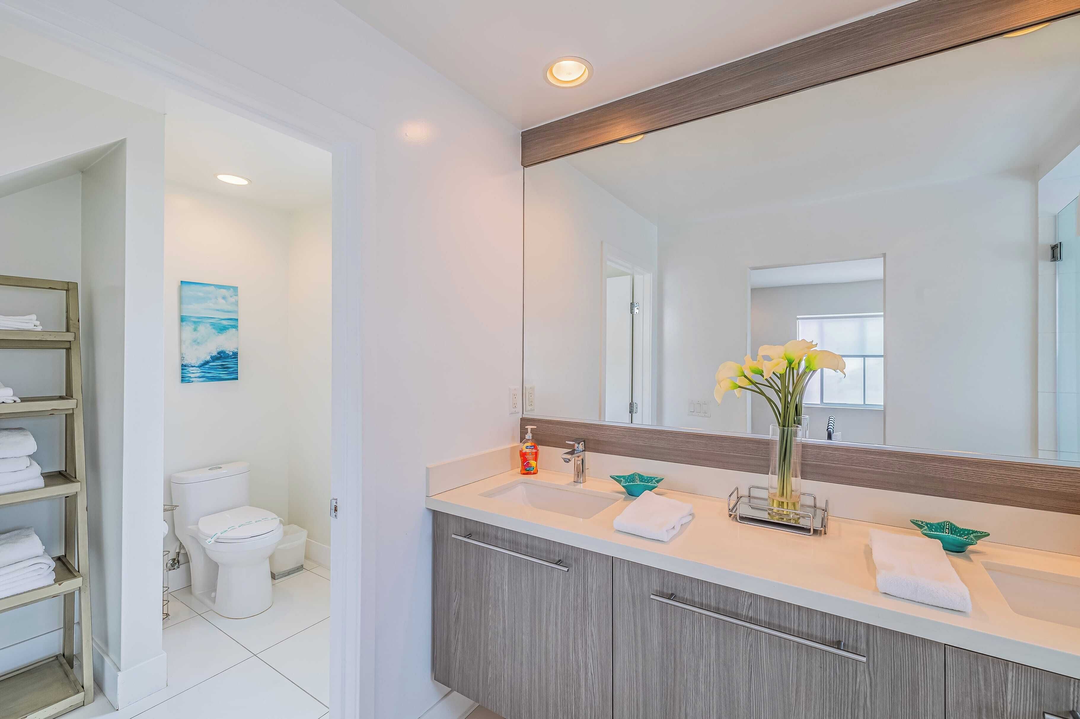 A modern bathroom with a white sink, a mirror, and a toilet. There is a shelf with folded towels and a painting on the wall.