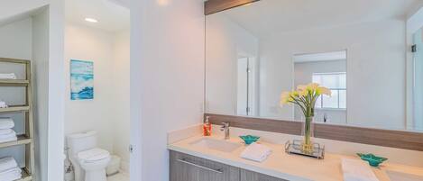 A modern bathroom with a white sink, a mirror, and a toilet. There is a shelf with folded towels and a painting on the wall.