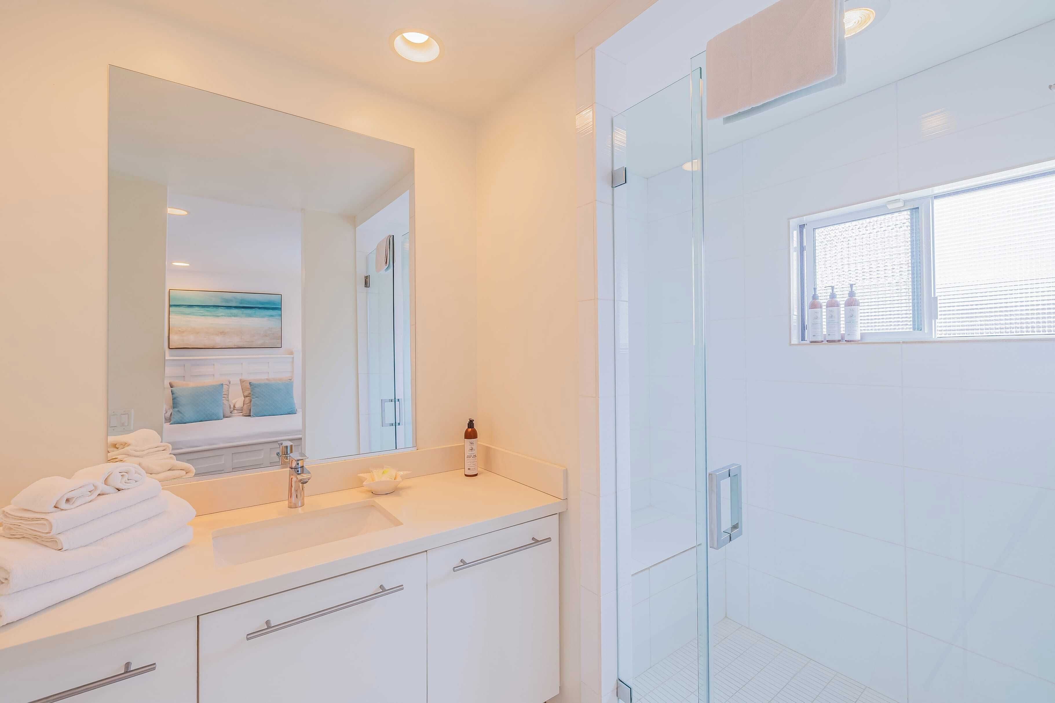 A modern bathroom with a large mirror, white tiles, a glass shower enclosure, and a window with bottles on the windowsill.