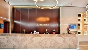 Ein Hotelrezeptionsschalter mit Marmorplatte, eine dunkle Wand mit dem Hotelnamen und ein Empfangsbereich mit Fernseher und Dekoration.