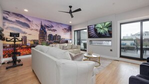 A modern living room with a cityscape mural, a ceiling fan, a large TV, and a view through large windows.