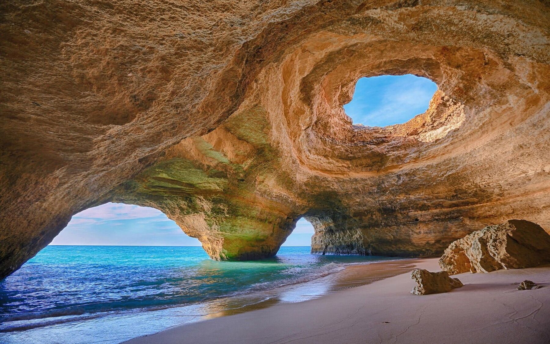 Formação rochosa natural com uma abertura circular, uma praia de areia e o oceano.