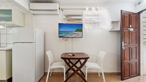 A compact kitchen with a refrigerator, a dining table with chairs, a TV mounted on the wall, and a wooden door. - Studio 1-Anthurium - A 7min de la plage (Les Trois-Îlets)