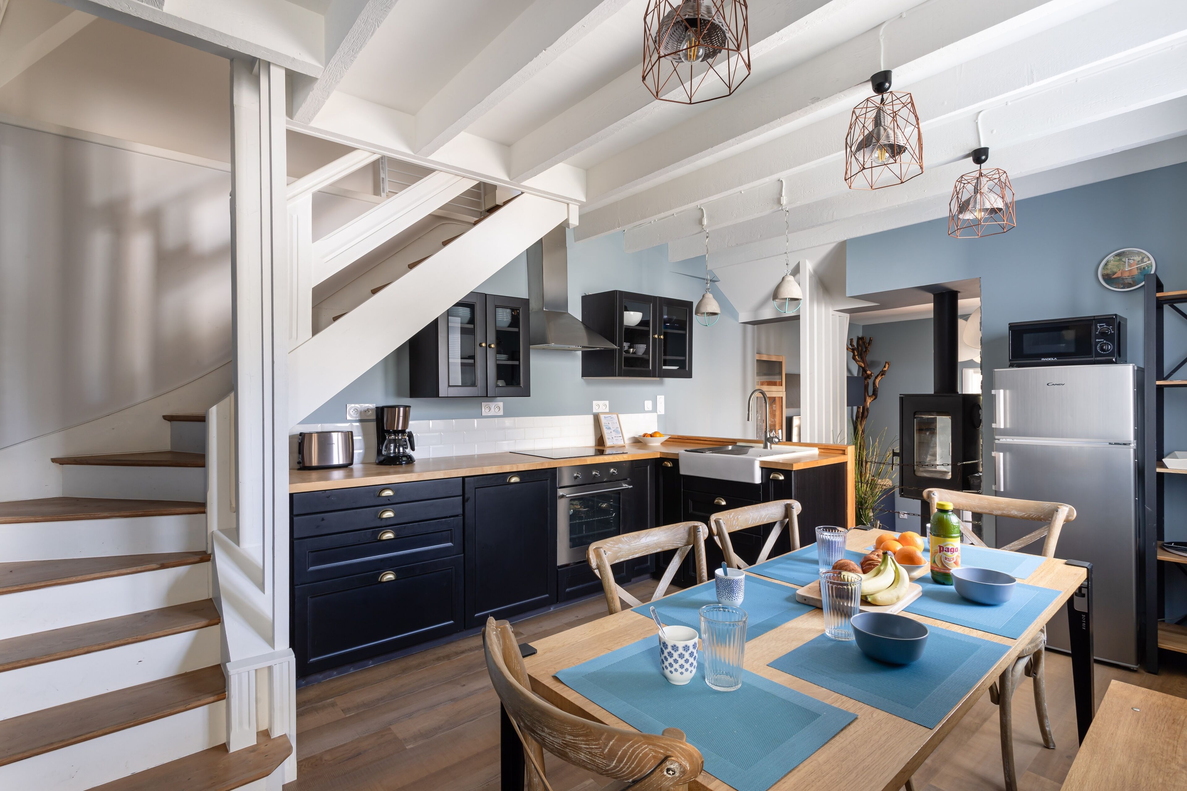 Une cuisine moderne avec des meubles de couleur sombre, un escalier en bois et un coin repas agrémenté de sets de table bleus.