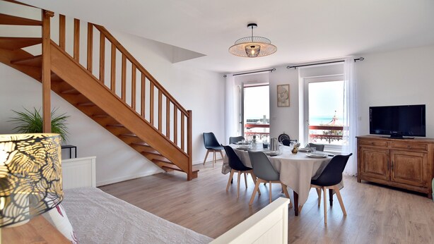 A modern living room with a wooden staircase, a dining area with a table and chairs, a television, and a view of the sea through large windows.