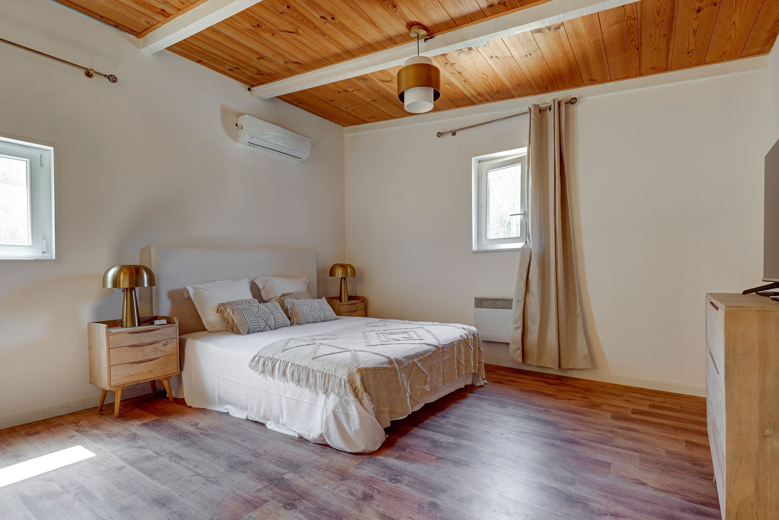 Quarto com teto de madeira, cama com colcha branca, duas luminárias de cabeceira e janela com cortinas.