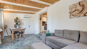 Ein Wohnzimmer mit grauem Sofa, Esstisch aus Holz und Stühlen. Eine Treppe führt zu einer Küchenzeile.