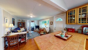 A cozy living room with a wooden coffee table, a sofa, and a bookshelf. There is a dining table with a tray of food and a red cooler. - "Charming 2-Bedroom Home in Narragansett with Private Beach Access and WiFi" (Narragansett)