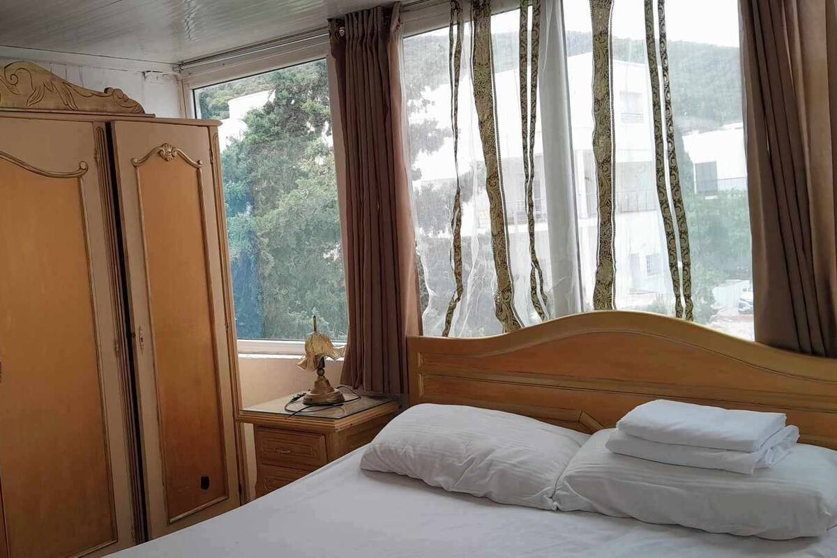 A bedroom with a wooden wardrobe, a bedside table with a lamp, and a bed with pillows.