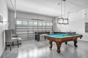 Uma mesa de bilhar com superfície de feltro verde, uma mesa de futebol de mesa em preto e branco, e uma área de estar com duas cadeiras.