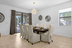 Uma sala de jantar com uma mesa grande posta para seis pessoas, seis cadeiras estofadas, um candelabro e duas placas decorativas penduradas na parede.