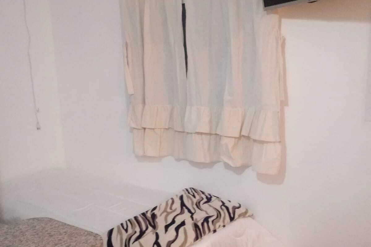 Una habitación pequeña y poco amueblada con una cama, un taburete pequeño, un reposapiés rojo y un televisor de pared.