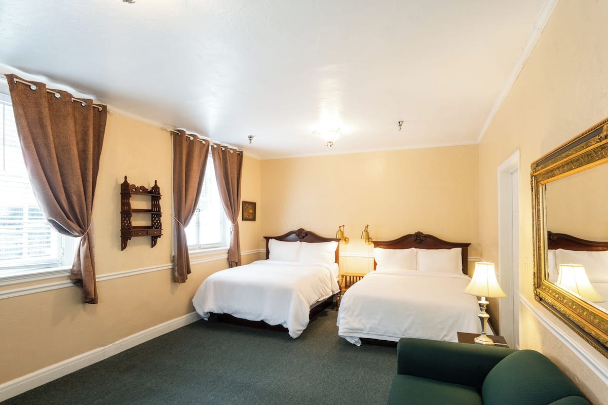 A hotel room with two beds, a green armchair, a large mirror, and a window with curtains.