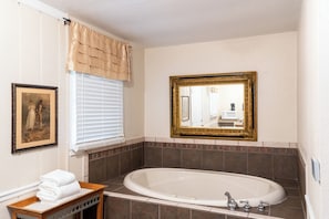 A bathroom with a bathtub, a framed mirror, a window with blinds, and a framed picture on the wall.