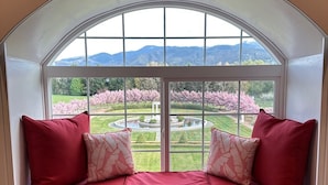 A window seat with red cushions and patterned pillows, overlooking a garden with pink flowering trees and a fountain. - Florence Room at Chateau Herbe (Ashland)