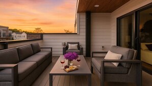 A rooftop terrace with a wooden deck, grey sofas, a table with a vase of flowers and wine glasses, and a city view at sunset. - Walk to West End | Rooftop Terrace + Deck (Nashville)