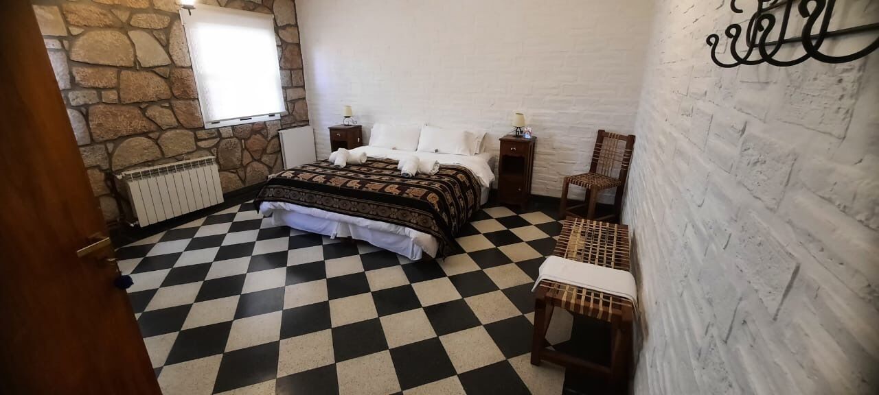Un dormitorio con piso a cuadros, una cama con colcha estampada, dos mesitas de noche con lámparas, un radiador y una ventana con persianas.