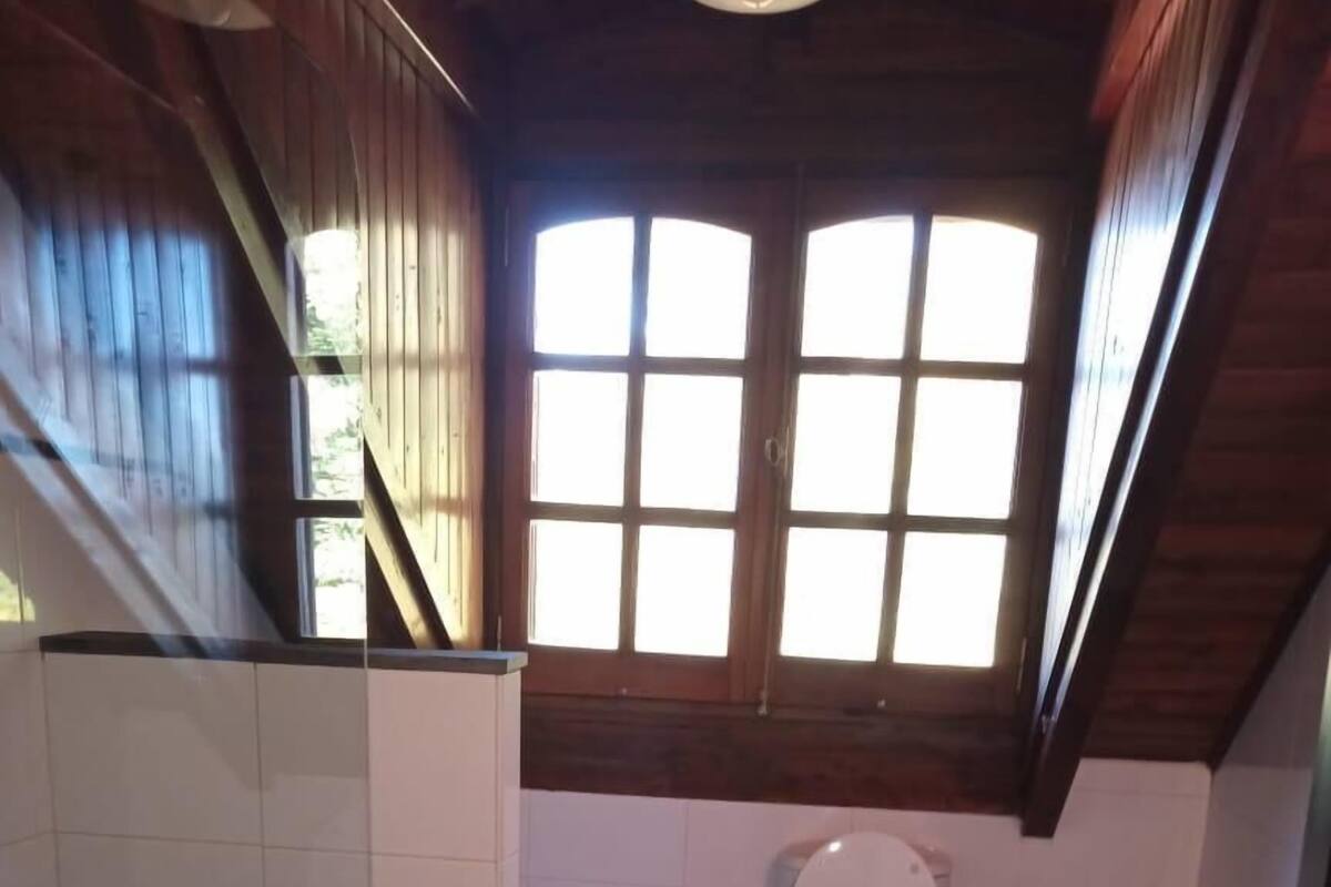 Een kleine badkamer met toilet, bidet en wastafel. Er is een raam boven het toilet en een houten trap aan de linkerkant.
