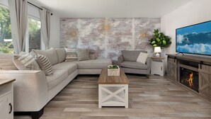 A modern living room with a sectional sofa, a wooden coffee table, a fireplace, and a large TV screen displaying a surfer. - 2 Condos, Minutes From Downtown, Parks, and Freeway! (Boise)