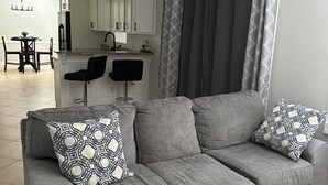 A living room with a grey sofa, patterned throw pillows, a ceiling fan, and a view into the kitchen.